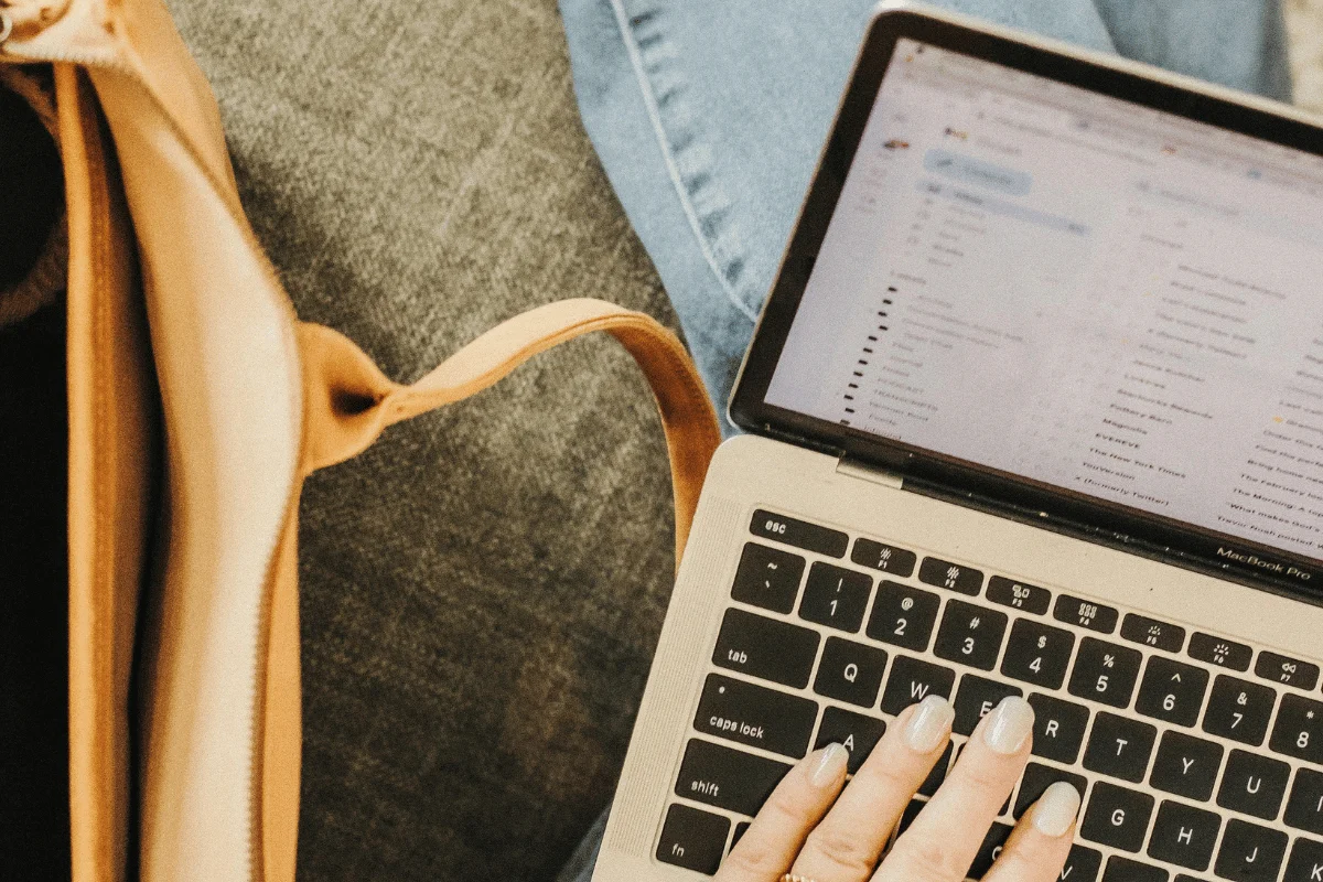 An individual's hand with manicured nails rests on a laptop keyboard, displaying a file directory alongside a beige handbag.