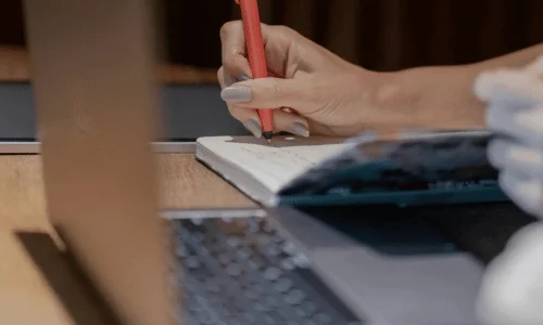 A hand with a red pencil writes in a notebook, with a laptop partially visible in the foreground on a wooden table.