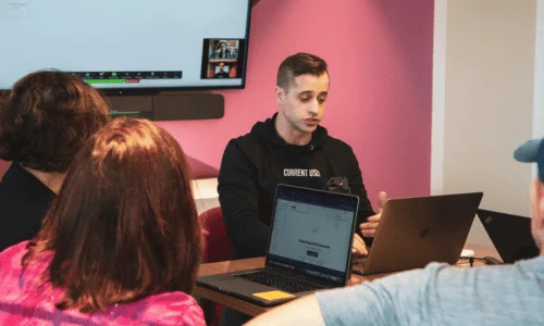 A group of people discuss ideas in a modern meeting room, with laptops open and a large screen displaying a video call in the background.