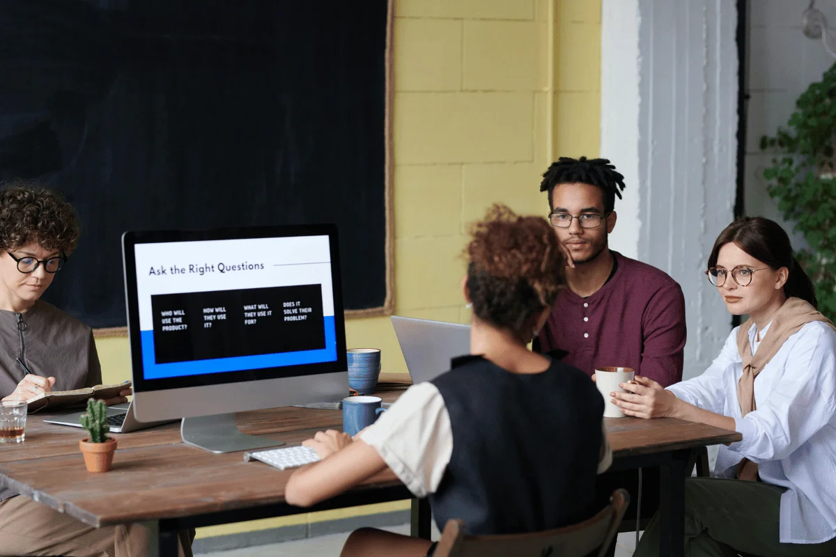 Dynamics 365 Customer Engagement Apps A group of four individuals seated at a table, engaging in discussion with a computer screen displaying strategic questions.