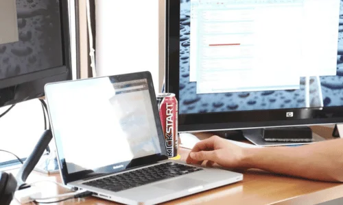A laptop and can of Kickstart energy drink sit on a wooden desk. A person's arm rests near a large monitor displaying blurred text, conveying a work setup.