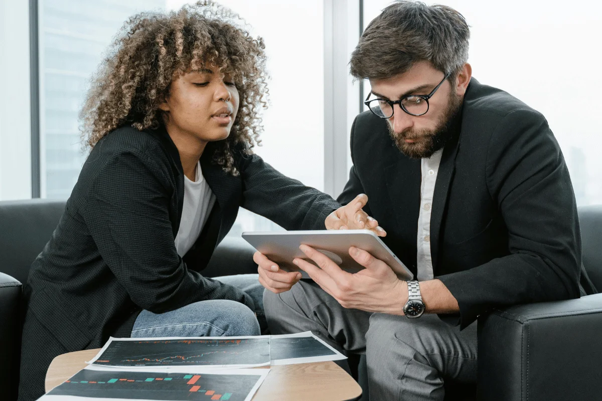 Two professionals collaboratively discussing a project on a tablet, with financial documents spread on a table in a modern office setting.