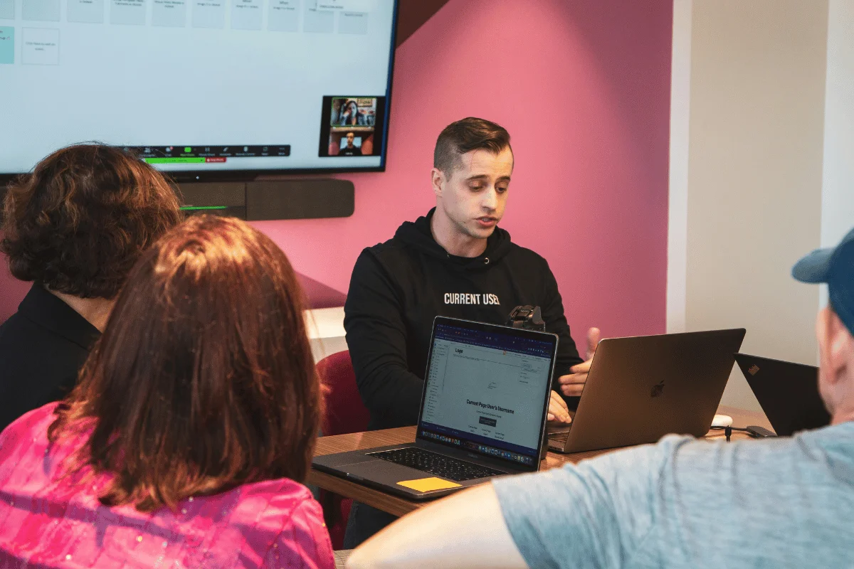 A man leads a meeting in a pink-walled room, speaking to three people around a table with laptops. A screen in the background displays slides.