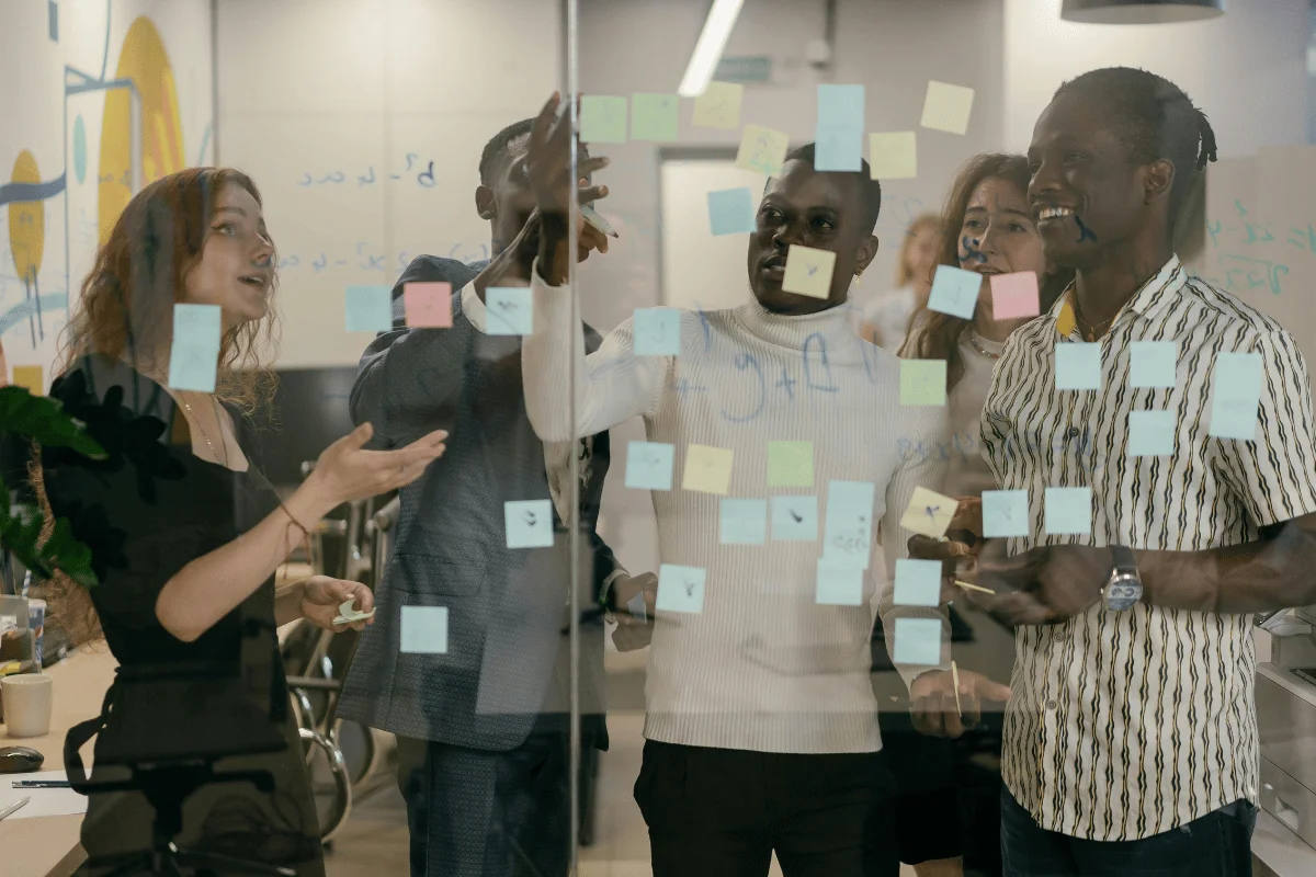 A diverse group of professionals collaborates in an office, discussing ideas while surrounded by colorful sticky notes on a glass wall.
