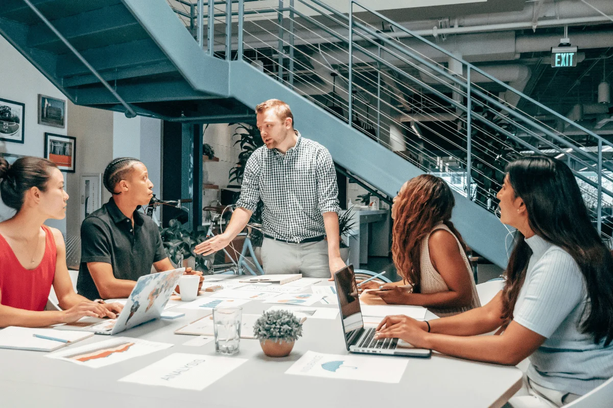 A team meeting in a modern office, with a man presenting and others engaged around a conference table filled with documents and laptops.