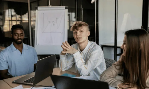A diverse group of three people sits at a conference table with laptops, listening intently. A whiteboard with a graph is in the background.