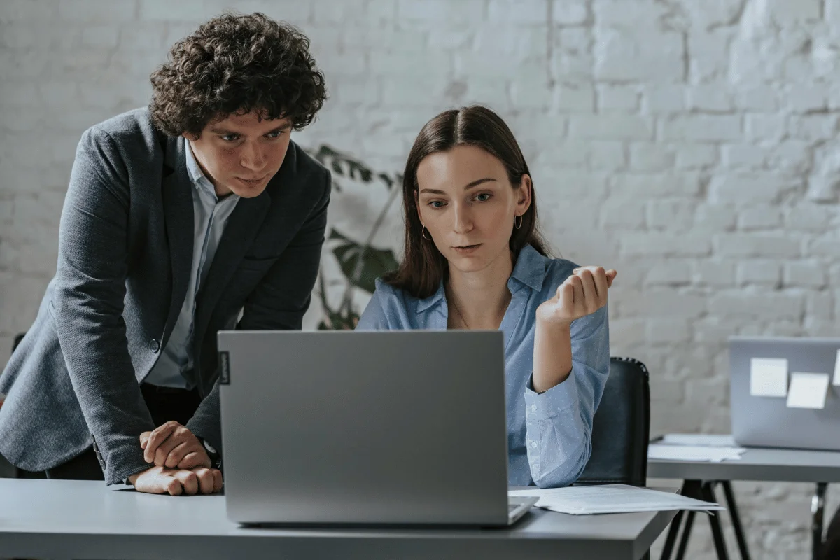 How to Set Up Microsoft Dynamics 365 Customer Engagement Two professionals discuss a project while seated at a desk with a laptop, surrounded by a modern office setting.