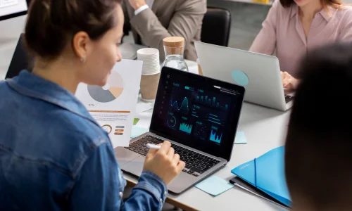 A woman in a denim shirt reviews data on a laptop with charts and graphs. Colleagues work alongside her at a table, creating a focused, collaborative atmosphere.