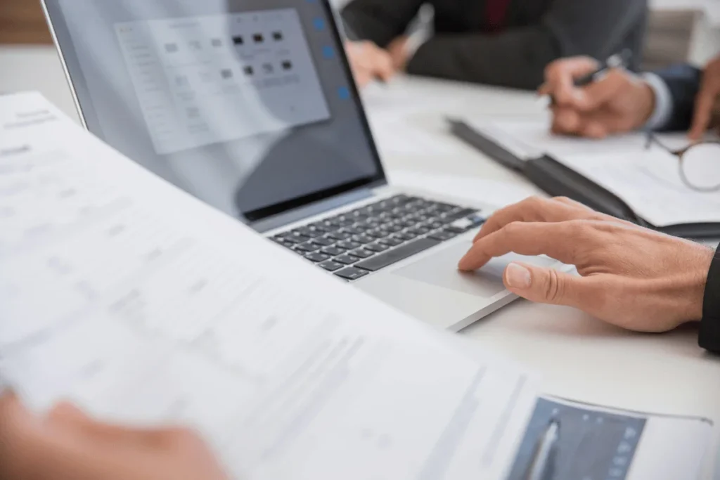 Business professional using laptop during meeting with printed reports.