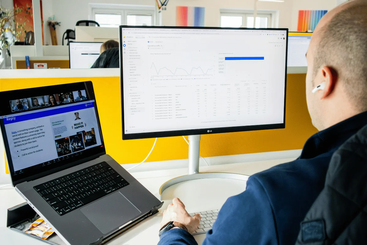 Man at desk analyzing data on dual monitors; one shows a webpage, the other displays charts and tables. Modern office setting, focused atmosphere.