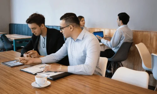 Three individuals hold a business meeting at a modern cafe, seated at a wooden table with laptops and a coffee cup.