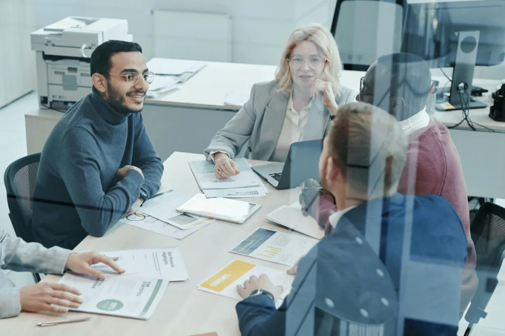A group of professionals engaged in a meeting, surrounded by documents and laptops on a conference table in a modern office setting.