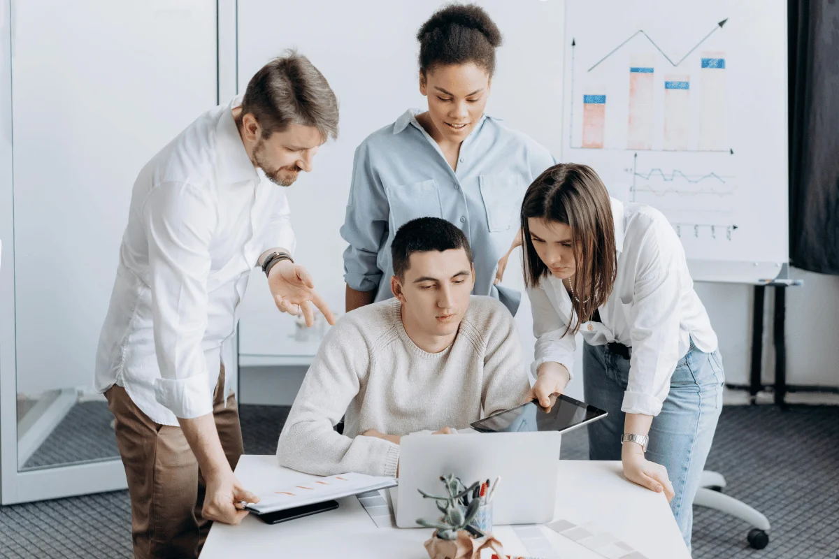 A team collaborates around a laptop, engaged in discussion, with charts on a flip chart in the background.