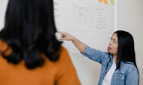 A woman in a denim jacket points at a whiteboard filled with diagrams while discussing with a colleague.
